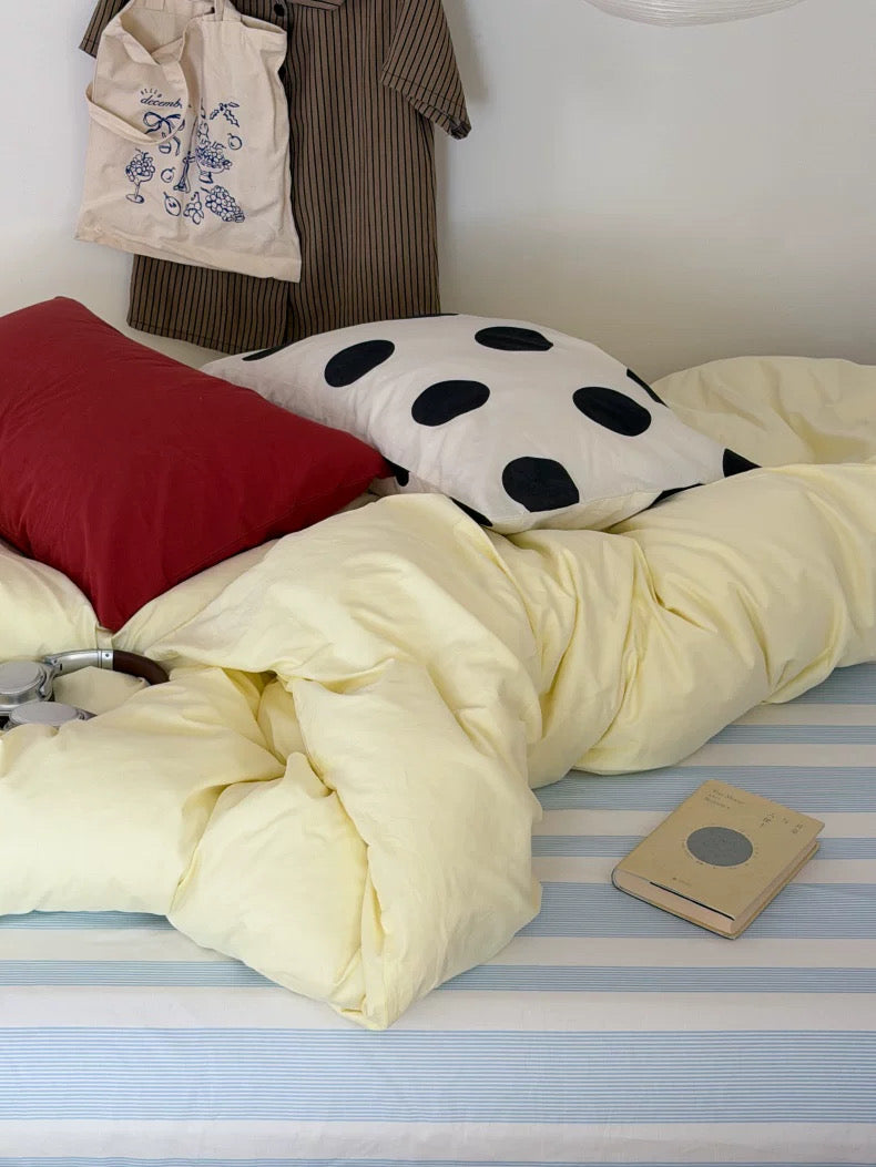 A cozy bed setup with pale yellow bedding, a red pillow, a white pillow with black polka dots, a blue-and-white striped sheet, and a small book on a light wall.