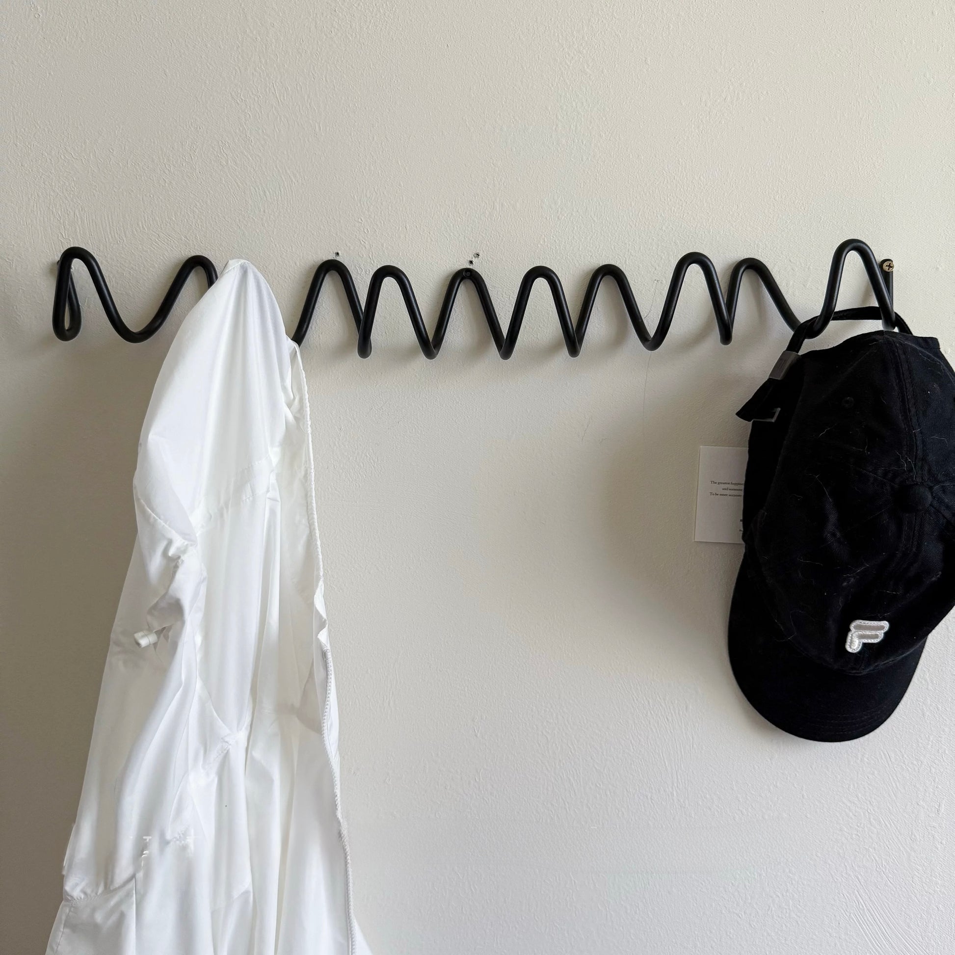 Black zigzag hook holding a white coat and a black cap on a light gray wall.