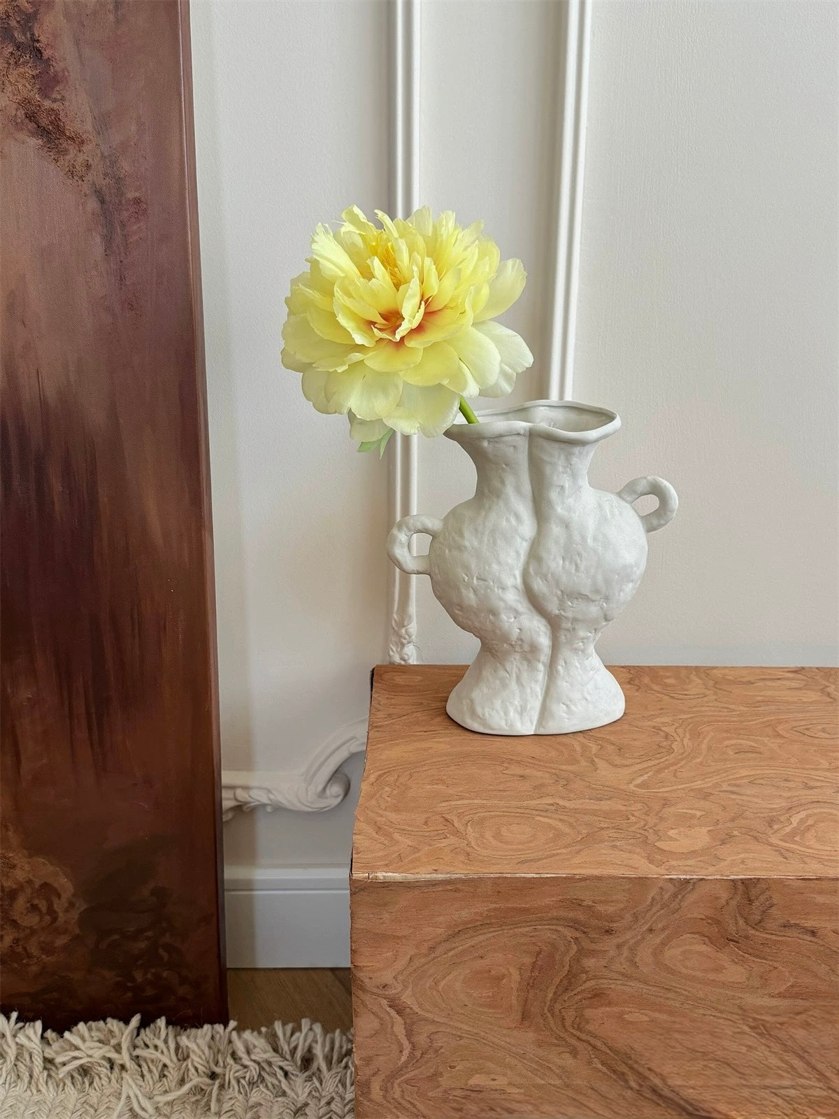 White vase with a yellow flower on a wooden surface next to a textured wall.
