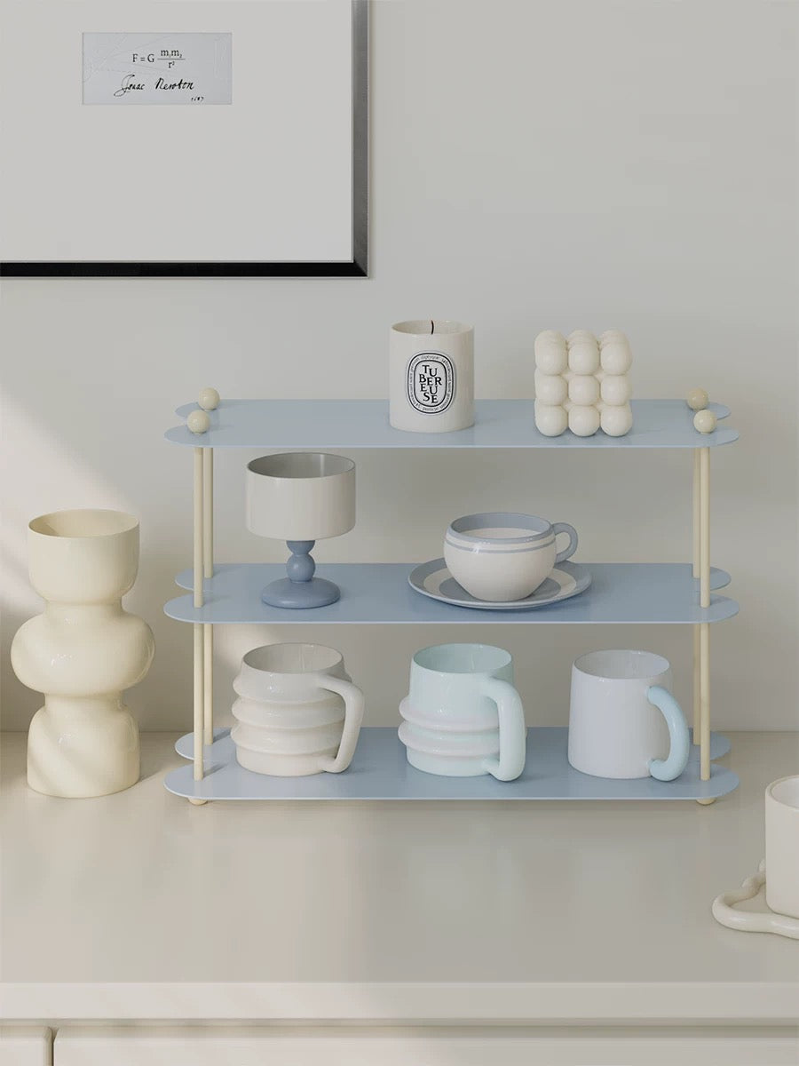 A blue mug display shelf with cream posts holds cups across three open levels, with candles on the top tier.