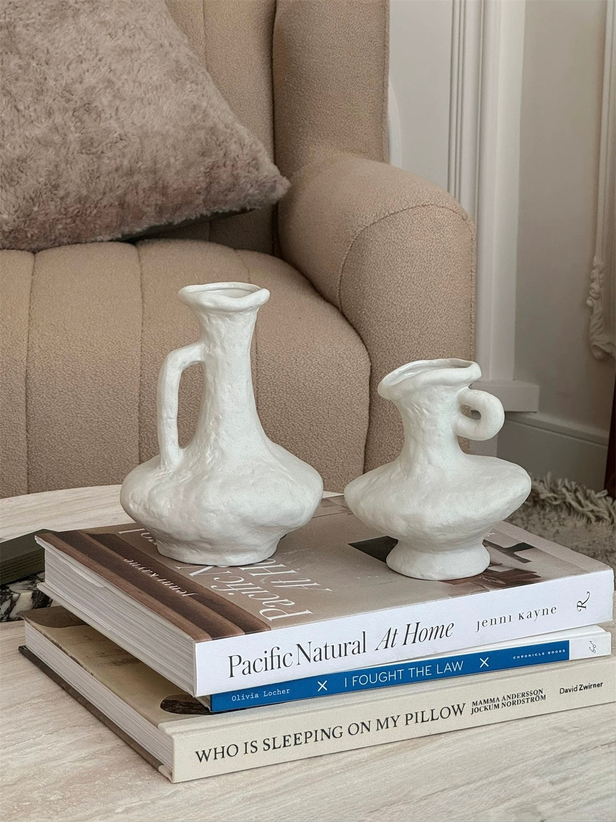 Two white ceramic vases on a stack of books with a beige sofa in the background