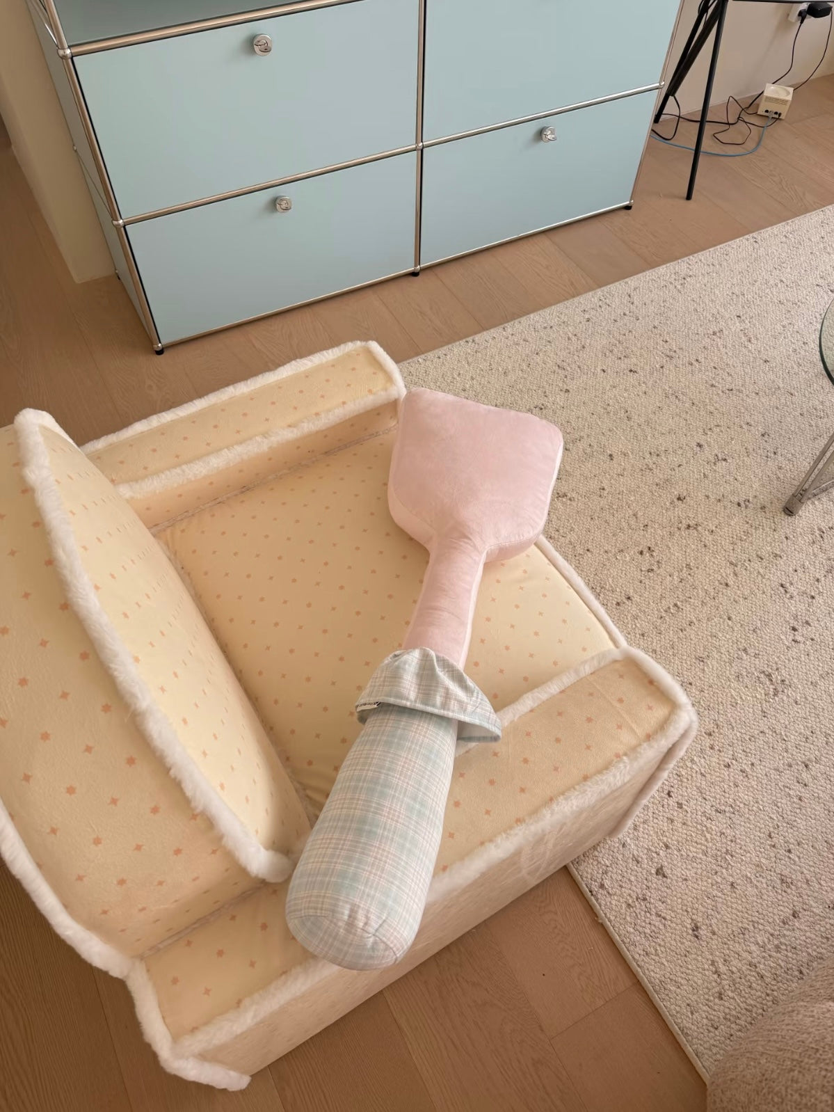 A pink spatula novelty pillow rests diagonally on a chair, showing a pale checked handle and gathered collar.