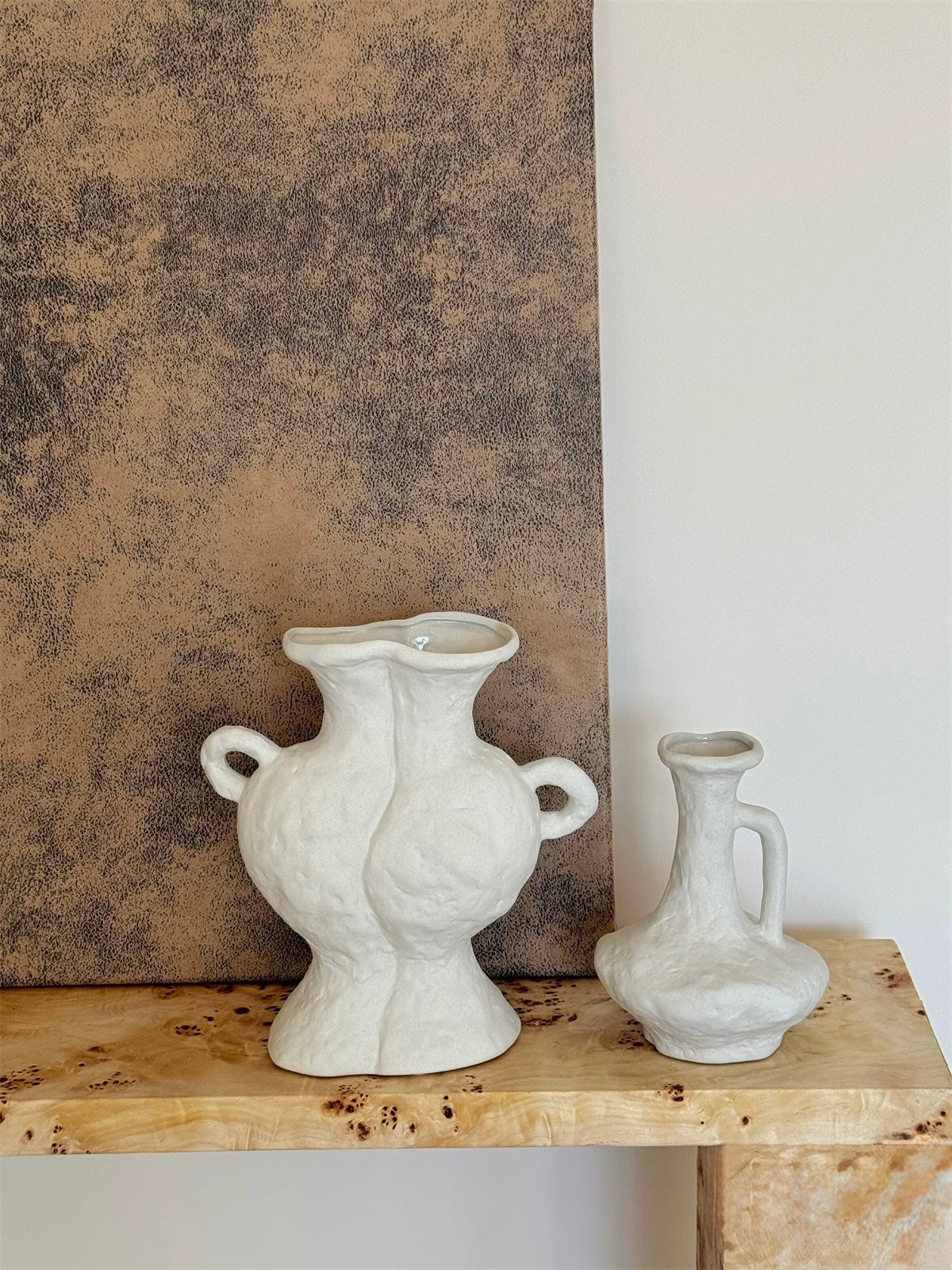 Two white ceramic vases on a wooden surface with a textured wall in the background.