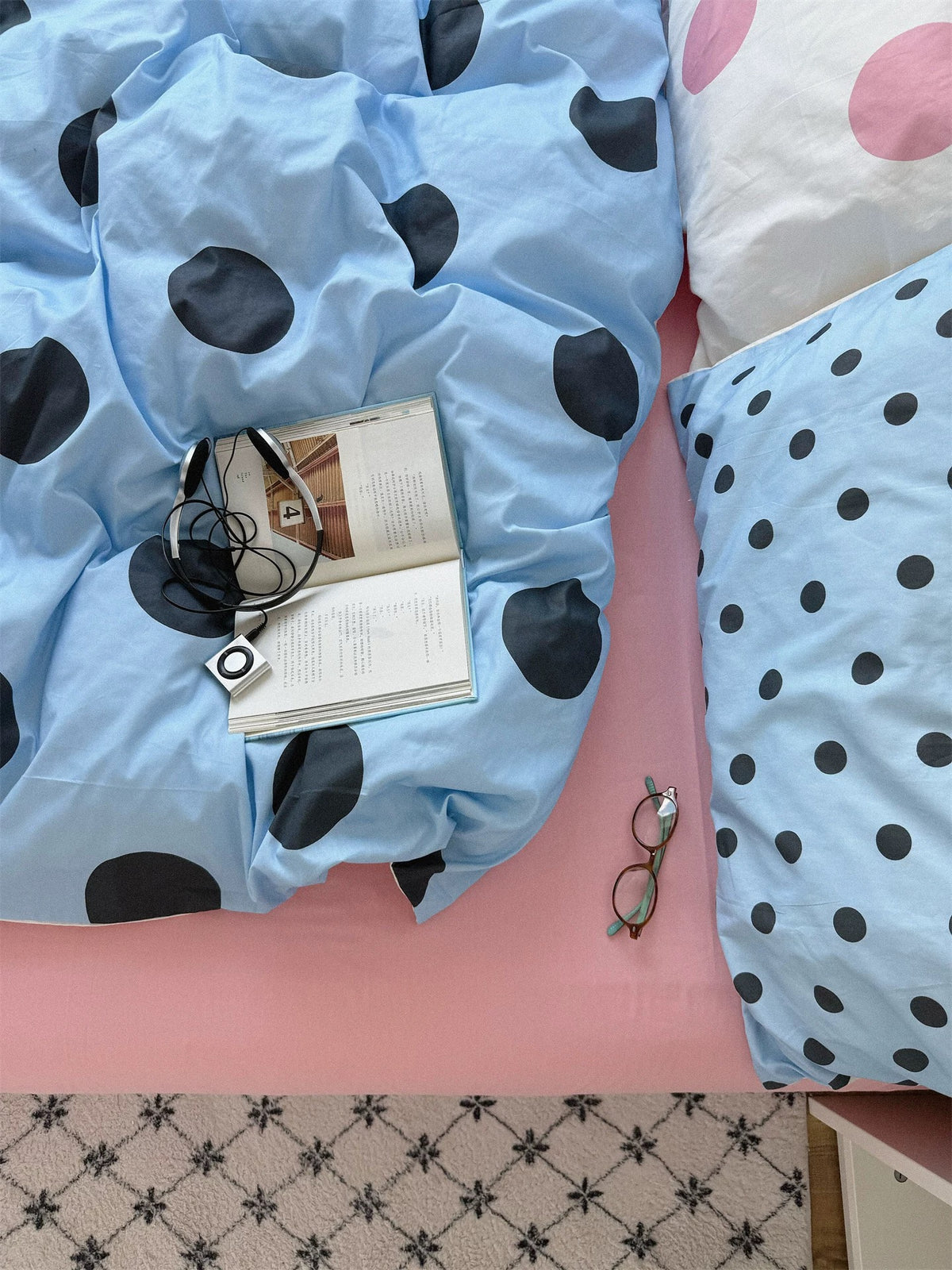 An overhead view shows oversized black dots on rumpled blue bedding beside a small dotted blue pillow and the pink sheet.