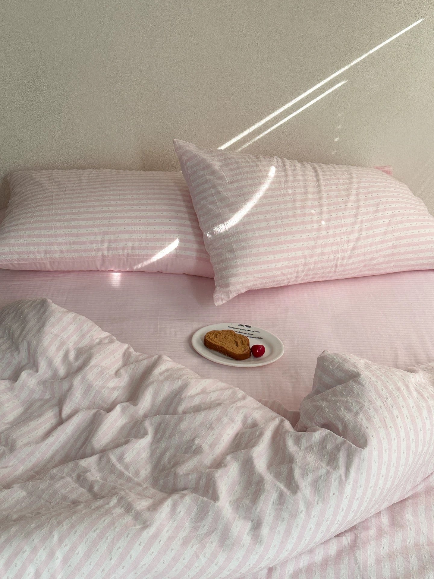 Pink and white striped bedding set on a bed, featuring two pillows, a matching duvet cover, and a small plate with toast and a cherry in the foreground.