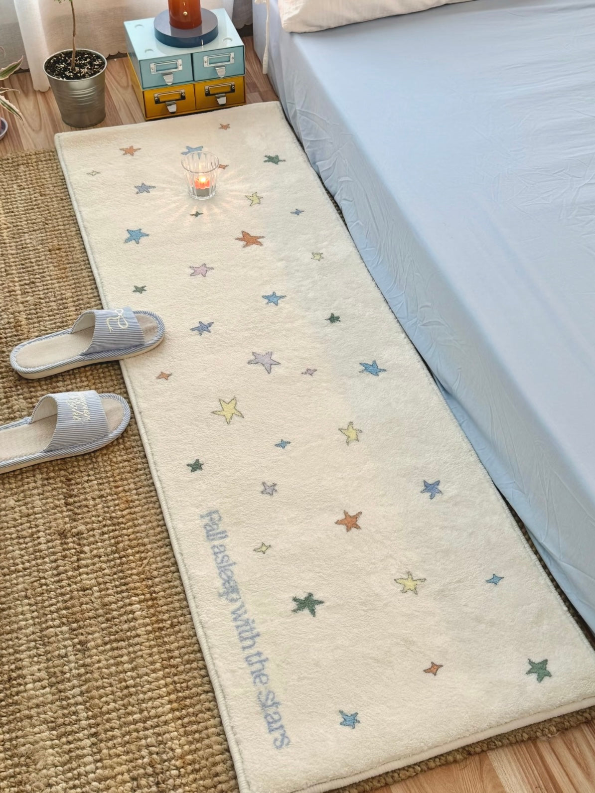 A pastel star runner rug shows mixed color motifs along a cream rectangle, viewed down the side of a bed.