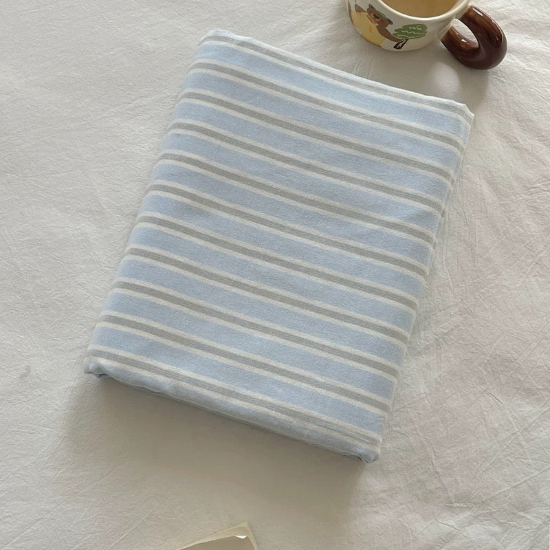 A folded striped fabric item, likely a blanket, towel, or pillowcase, in light blue and white, laid flat on a white textured fabric surface with a small decorative ceramic cup in the top right corner.