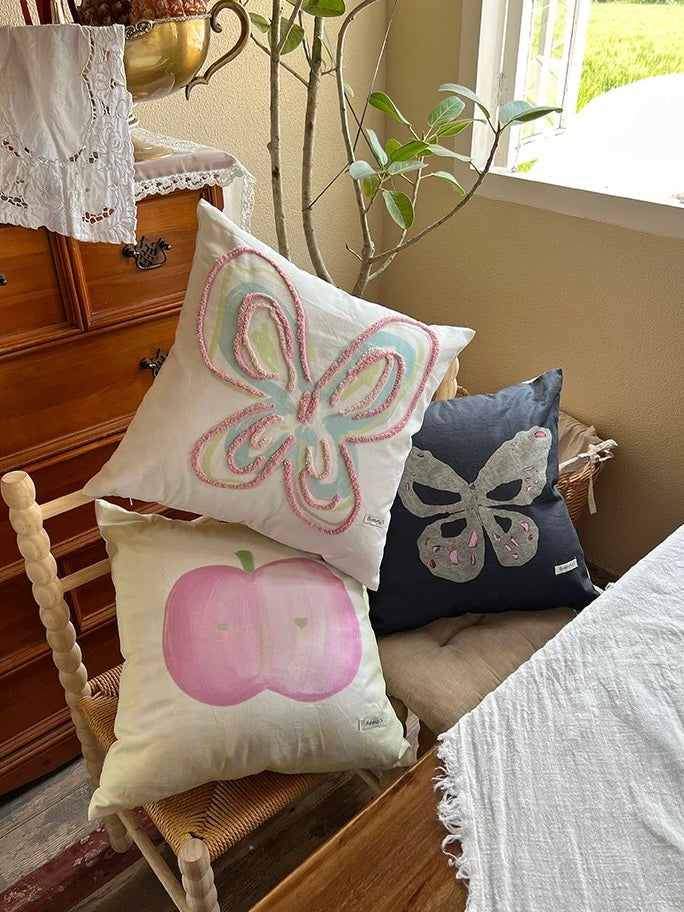 Pastel throw pillows with line butterfly, gray butterfly and apple designs stacked on a wooden chair