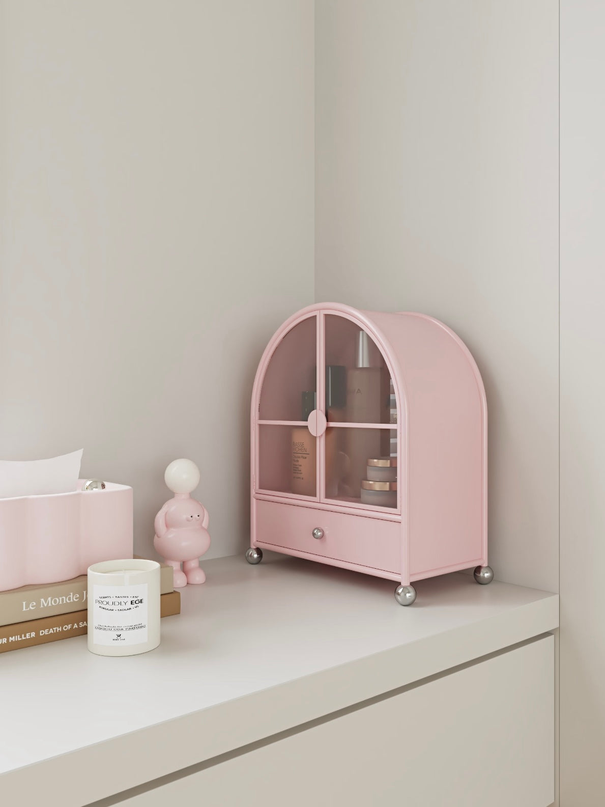 A pink arched cabinet is shown from the side on a dresser, with glass doors above its closed drawer.