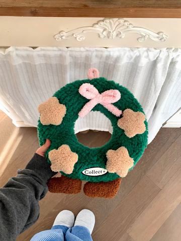 Green Christmas wreath pillow with pink bow and beige plush stars held in front of a white sofa