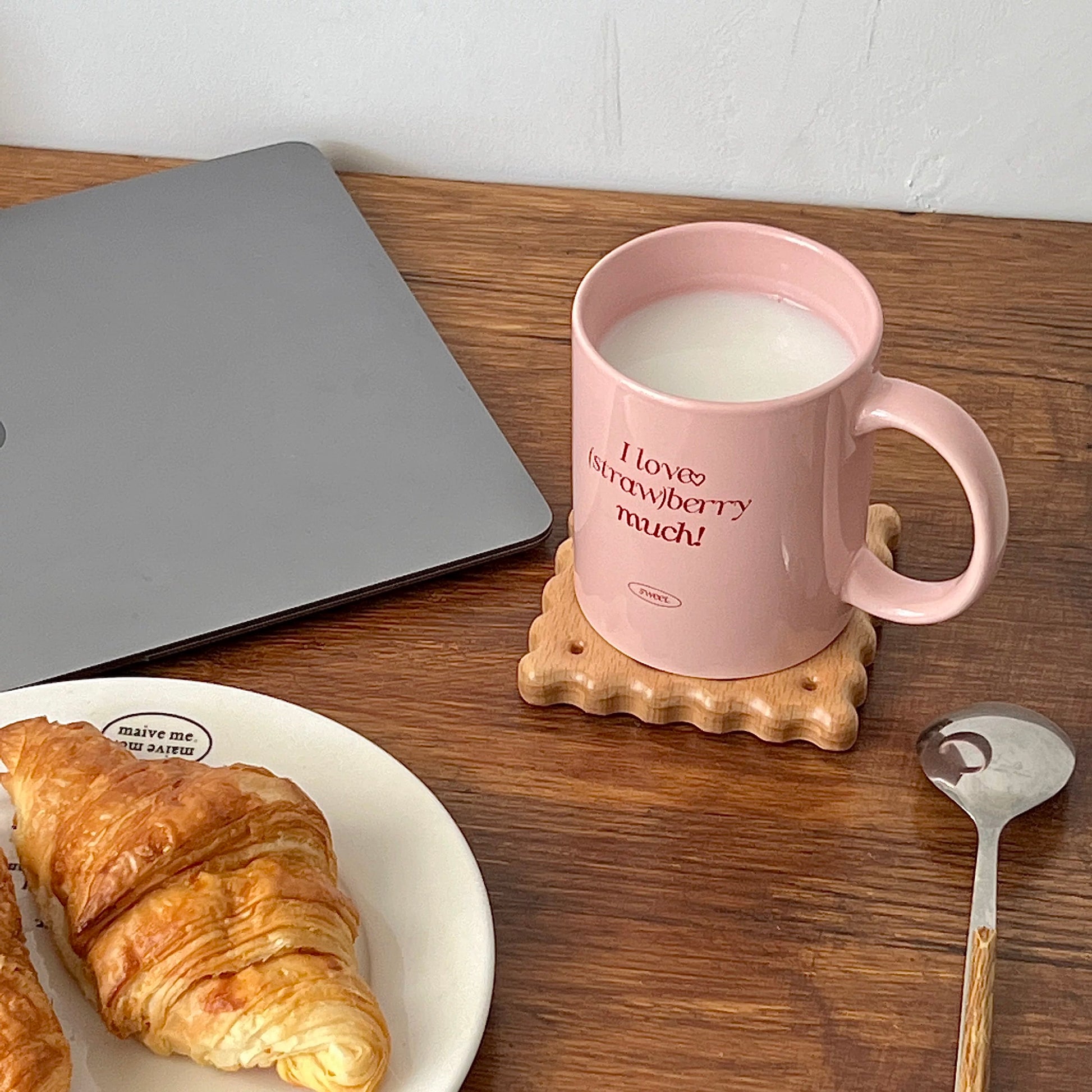 A pink ceramic mug filled with milk sits on a wooden coaster on a wooden table, surrounded by a laptop, a croissant on a plate, and a spoon. The mug has the text 'I love strawberry much!' printed on it.