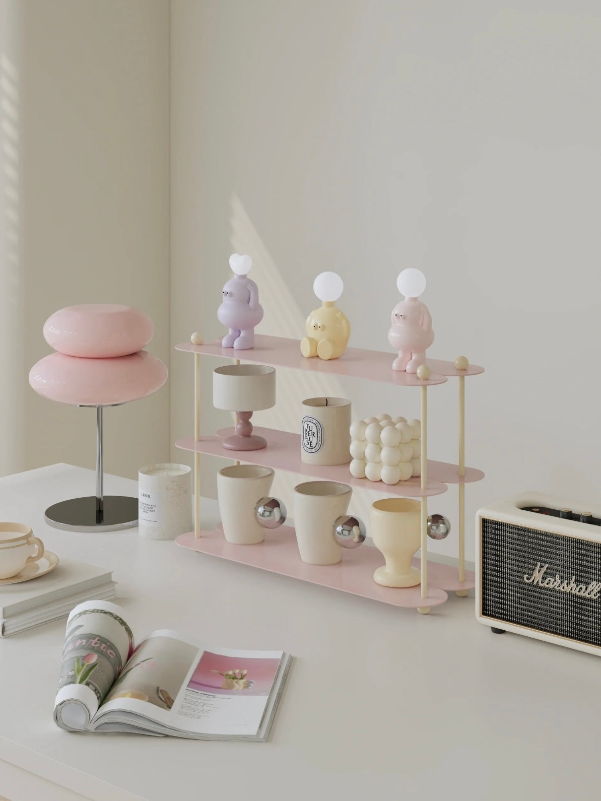 A pink display shelf with three open tiers sits on a desk beside a pink lamp and a cream speaker.
