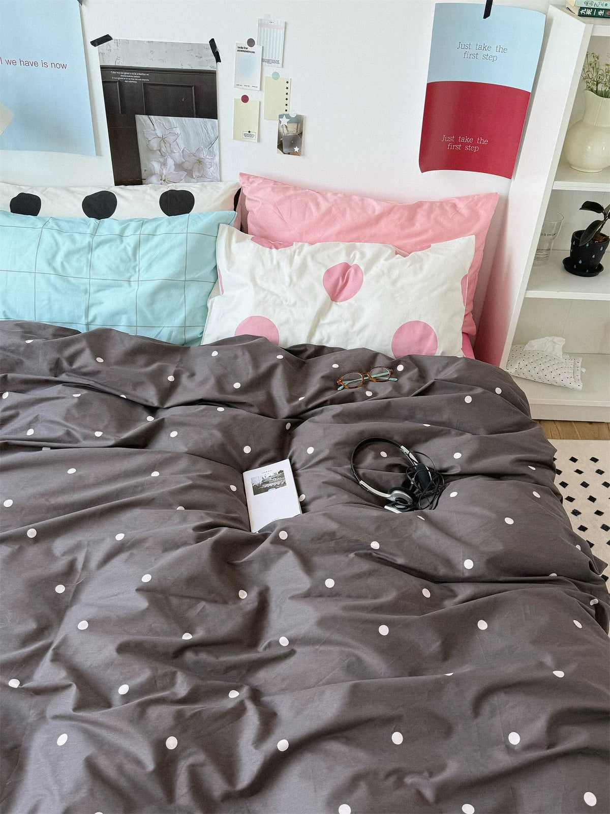 An overview of pink and gray bedding with a rumpled dotted cover beneath a group of solid, checked and dotted pillows.