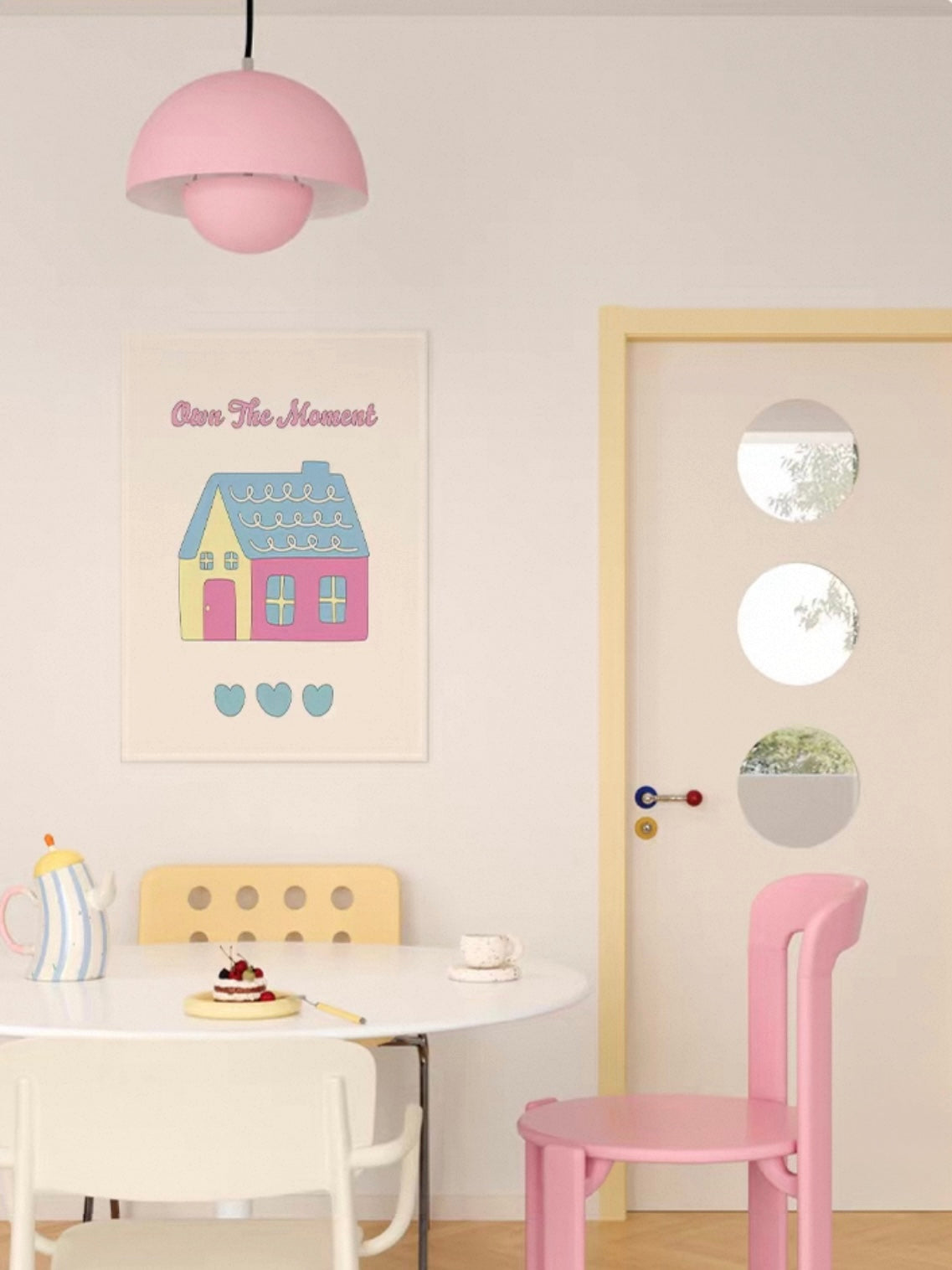 A pink house wall hanging with a blue roof appears above a small table beside a pink chair and pale door.