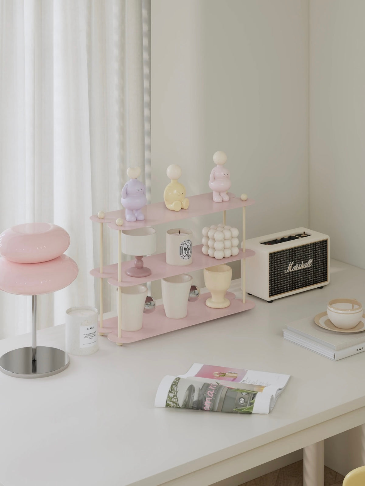 A pink iron shelf with cream posts is shown at an angle, with cups and candles displayed on its open tiers.