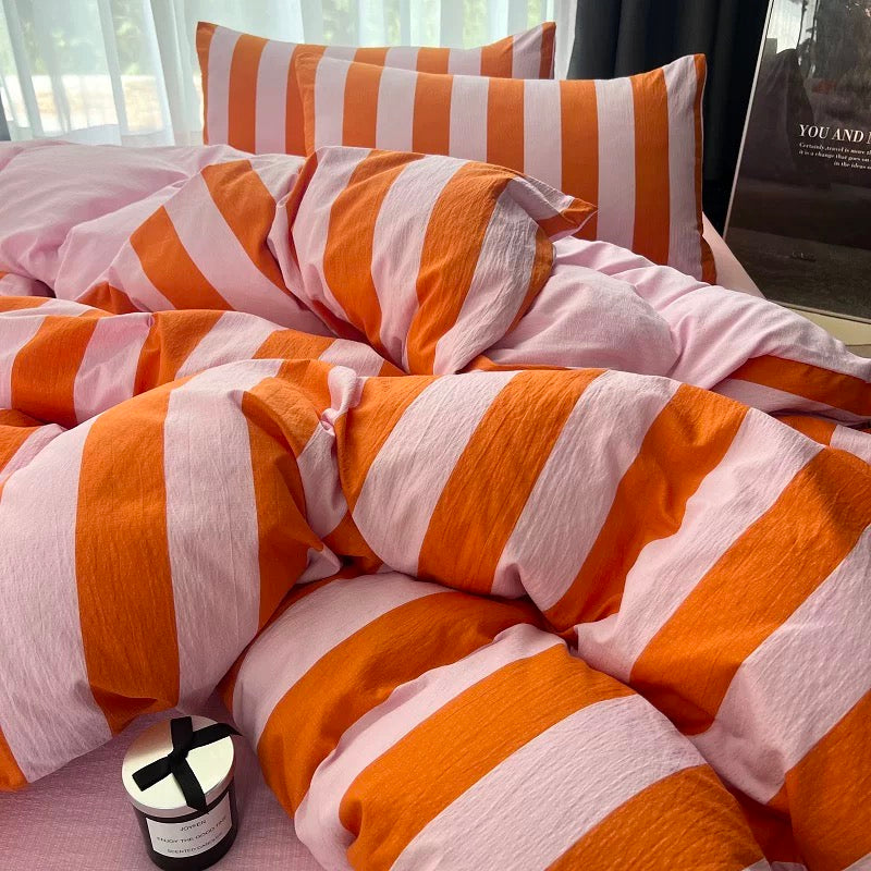 A bed made with a crinkled cotton bedding set featuring bold orange and white vertical stripes, styled in a bright bedroom setting with a candle and a framed print nearby.