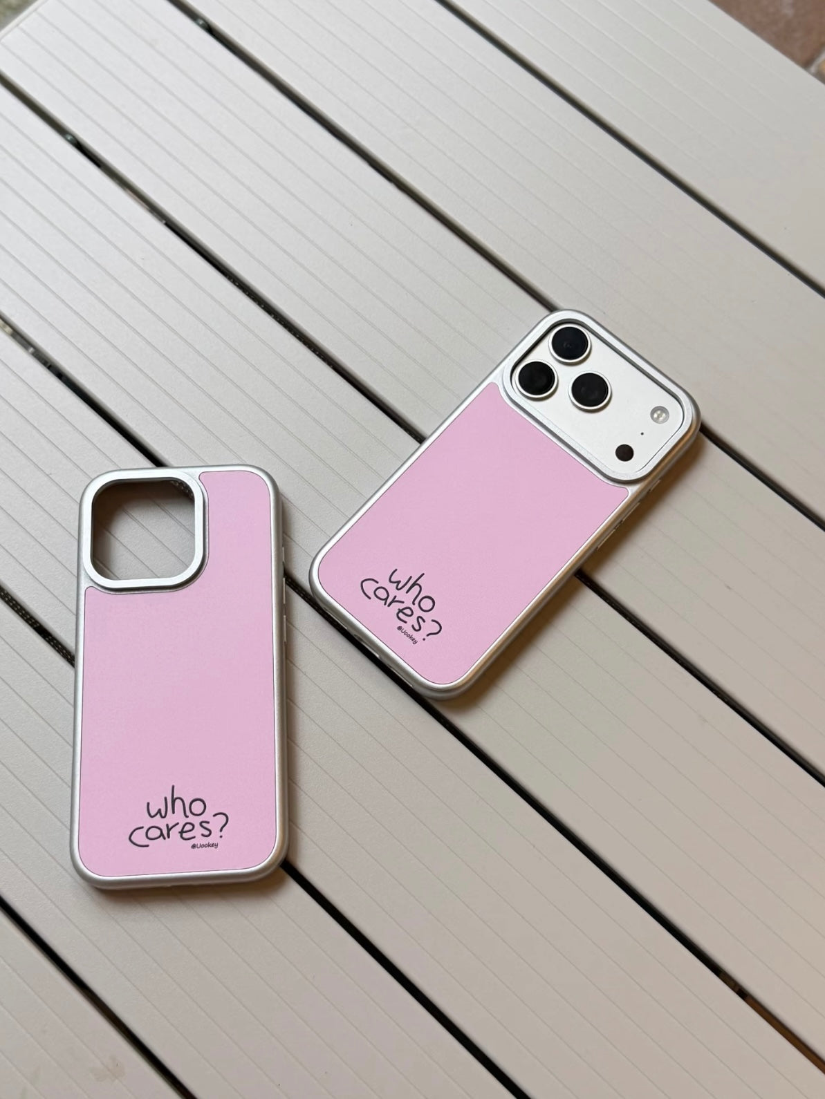 Two pink phone cases shown from the back have silver colored borders, black lettering and different camera areas.