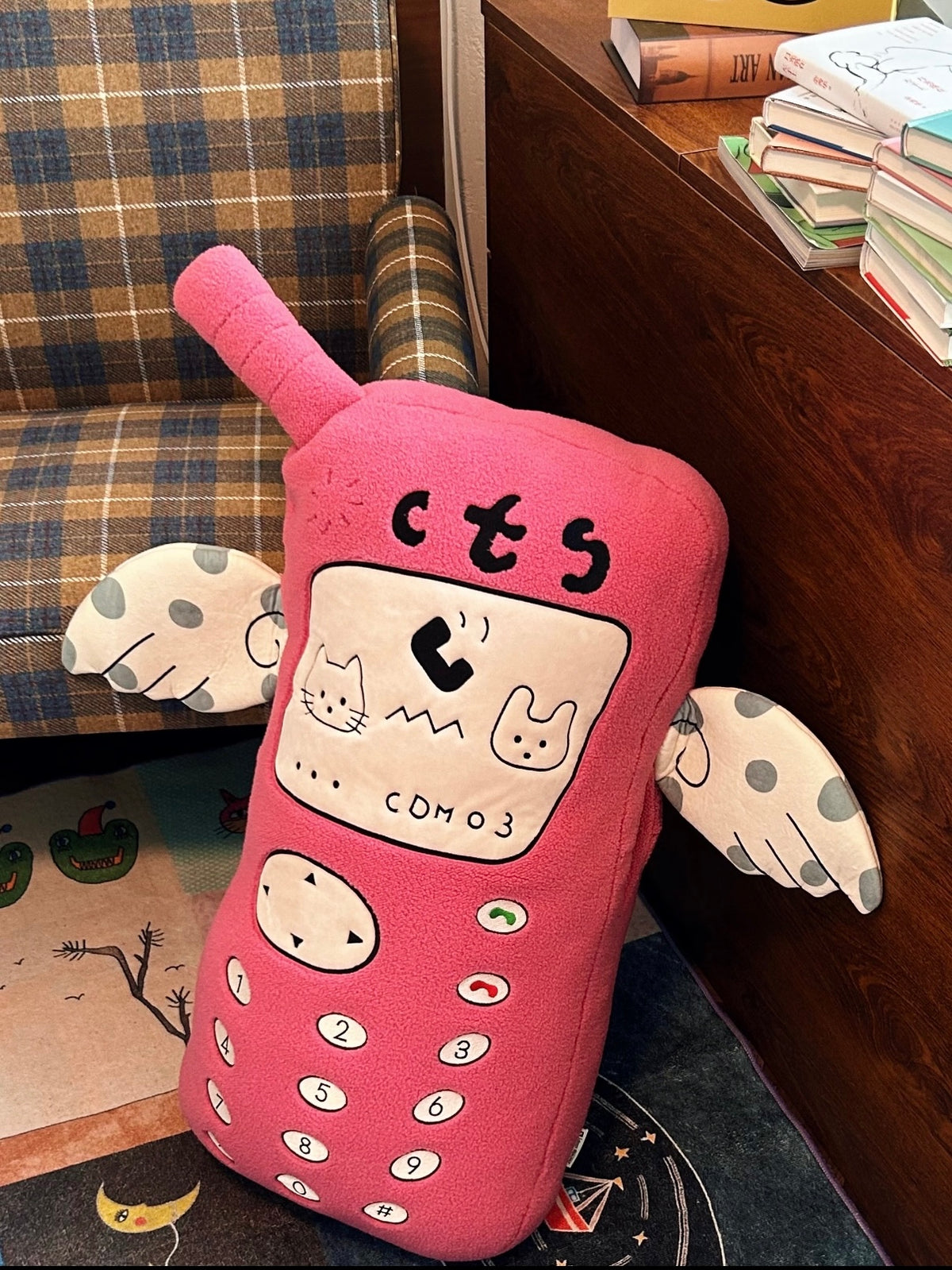 A pink phone pillow rests against a chair, with the keypad, cartoon screen faces and spotted arms visible.