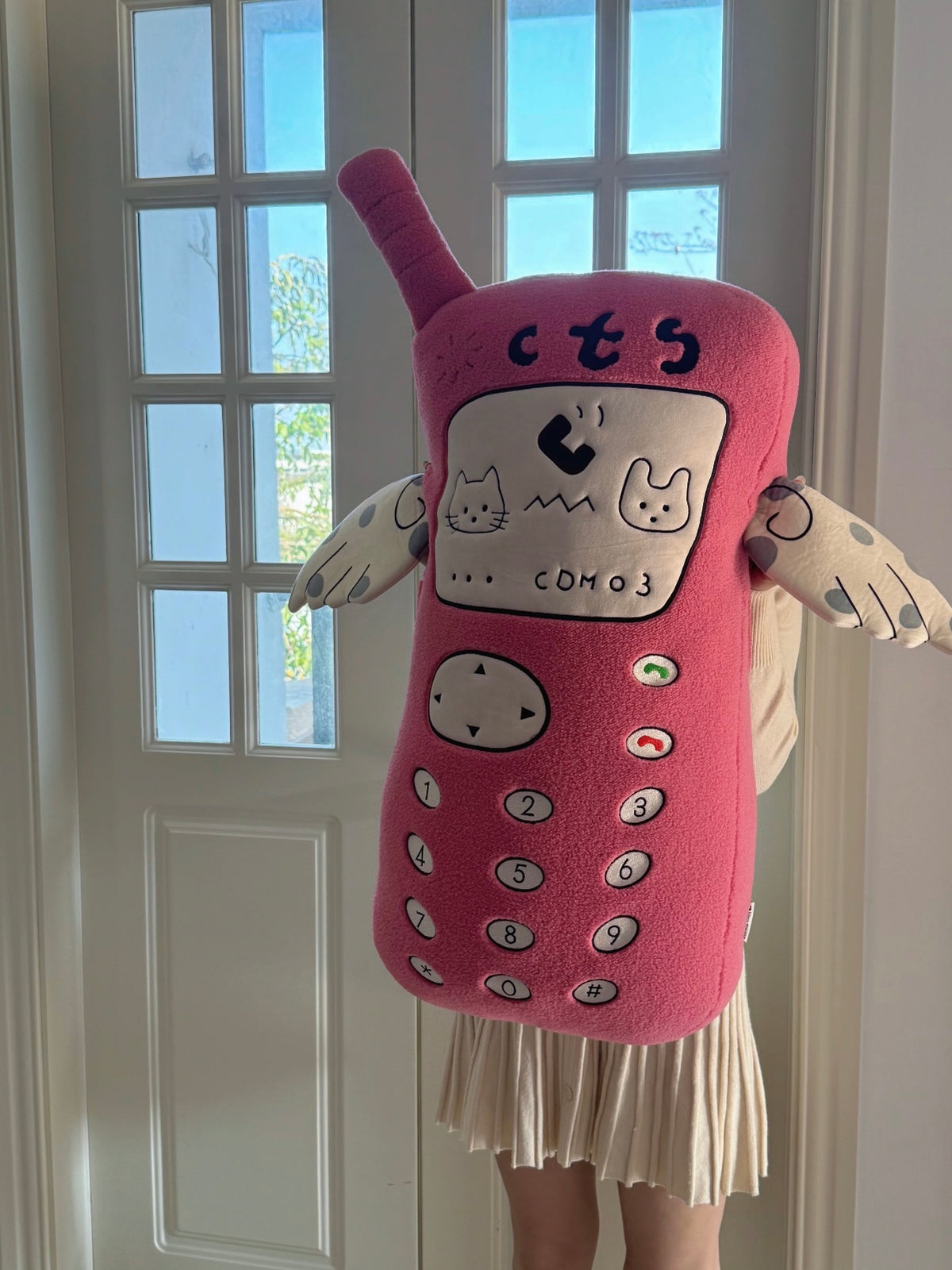 A person holds a pink telephone plush in front of them, with its antenna tilted above the embroidered keypad.