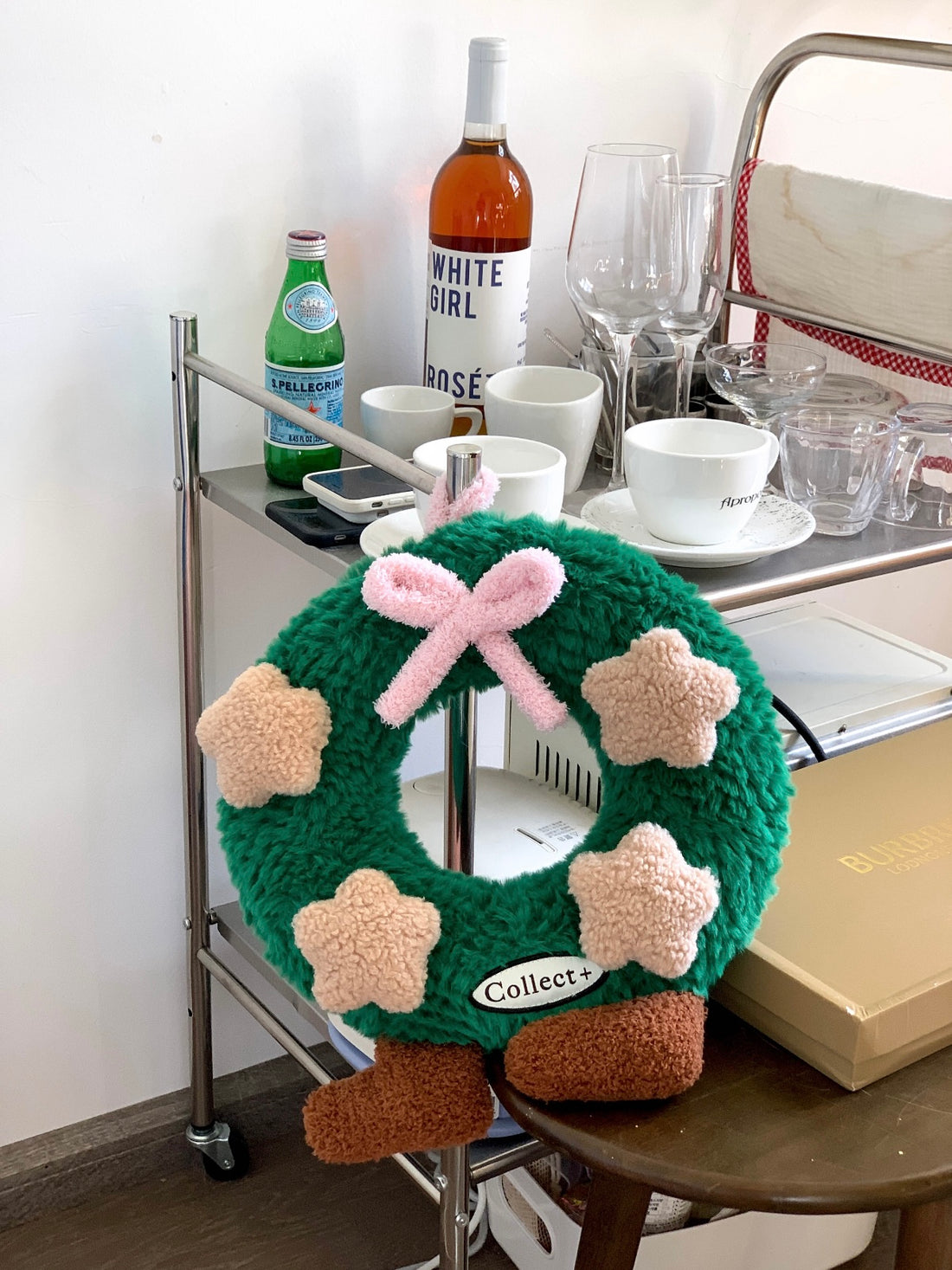 Green Christmas wreath pillow with pink bow and beige plush stars held in front of a white sofa