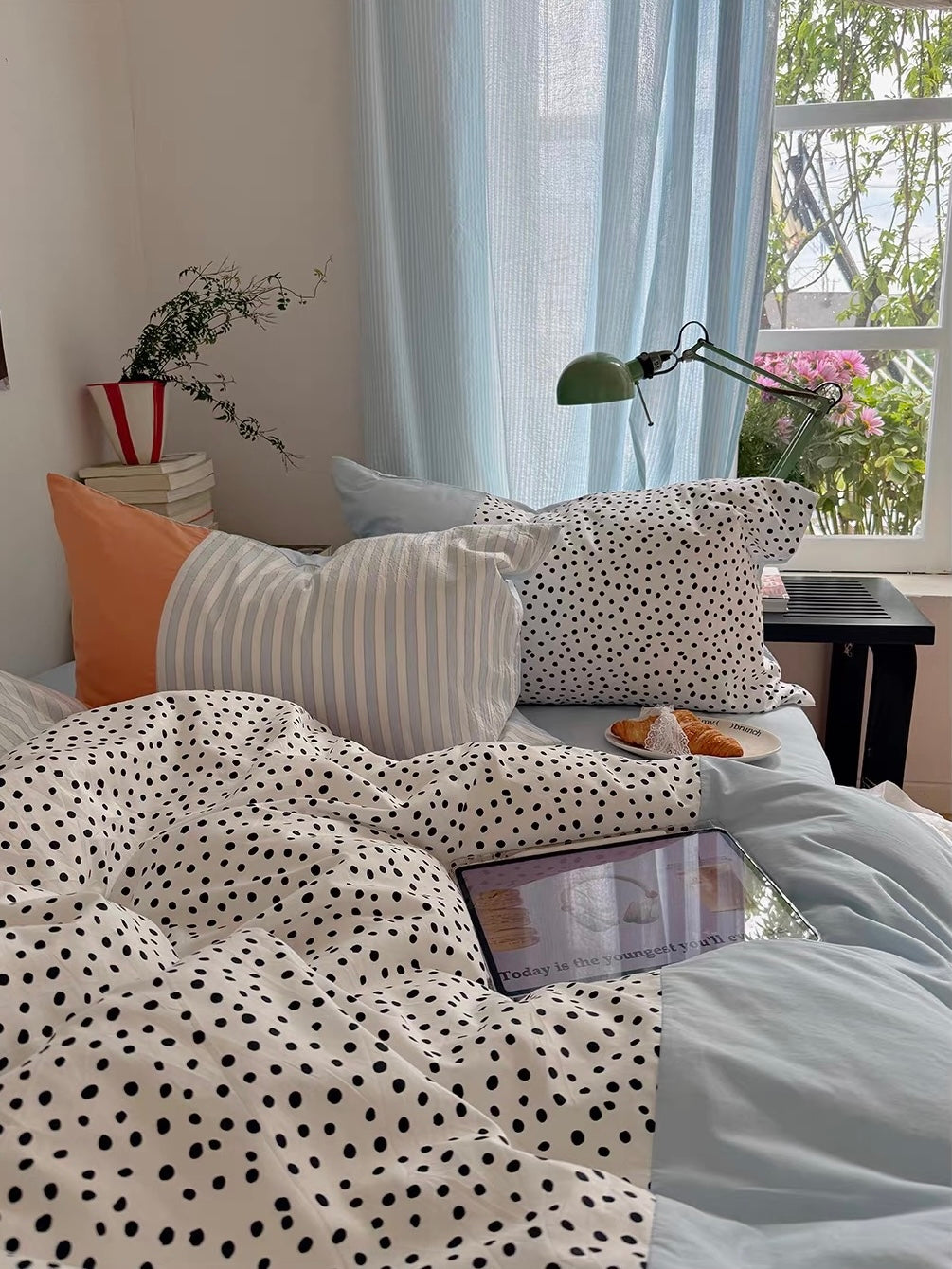 Polka dot bedding is pulled across a blue sheet beside a window, with striped, peach and dotted pillows.
