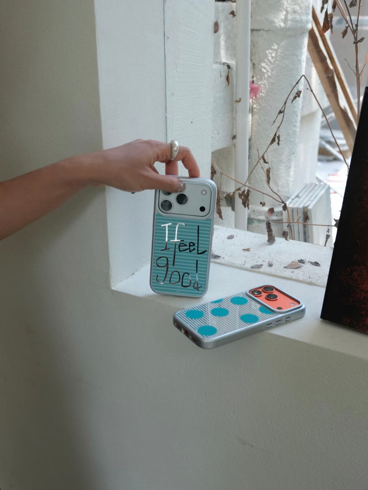 An aqua polka dot iPhone case lies beside the upright Feel Good design, with both silver borders visible.
