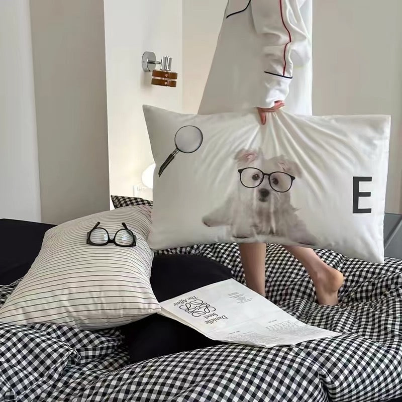 Person holding a pillow with a dog wearing glasses design, sitting on a bed with another pillow featuring glasses.