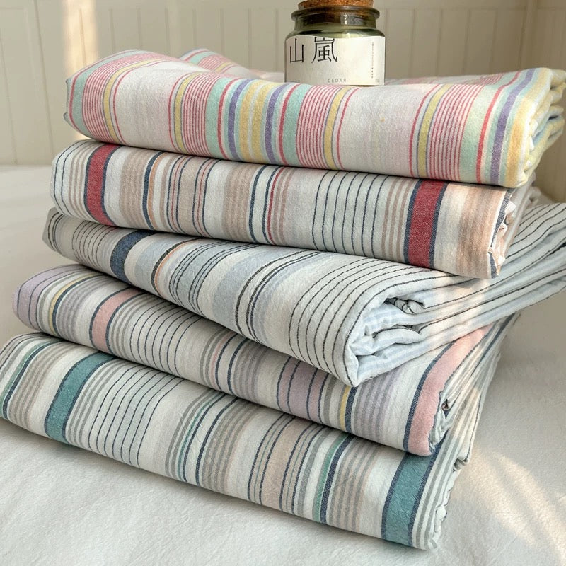 A stack of five folded striped textiles, likely towels, blankets, or sheets, in soft pastel colors including pink, blue, yellow, and green on a white base. A small glass jar with a white label sits on top.
