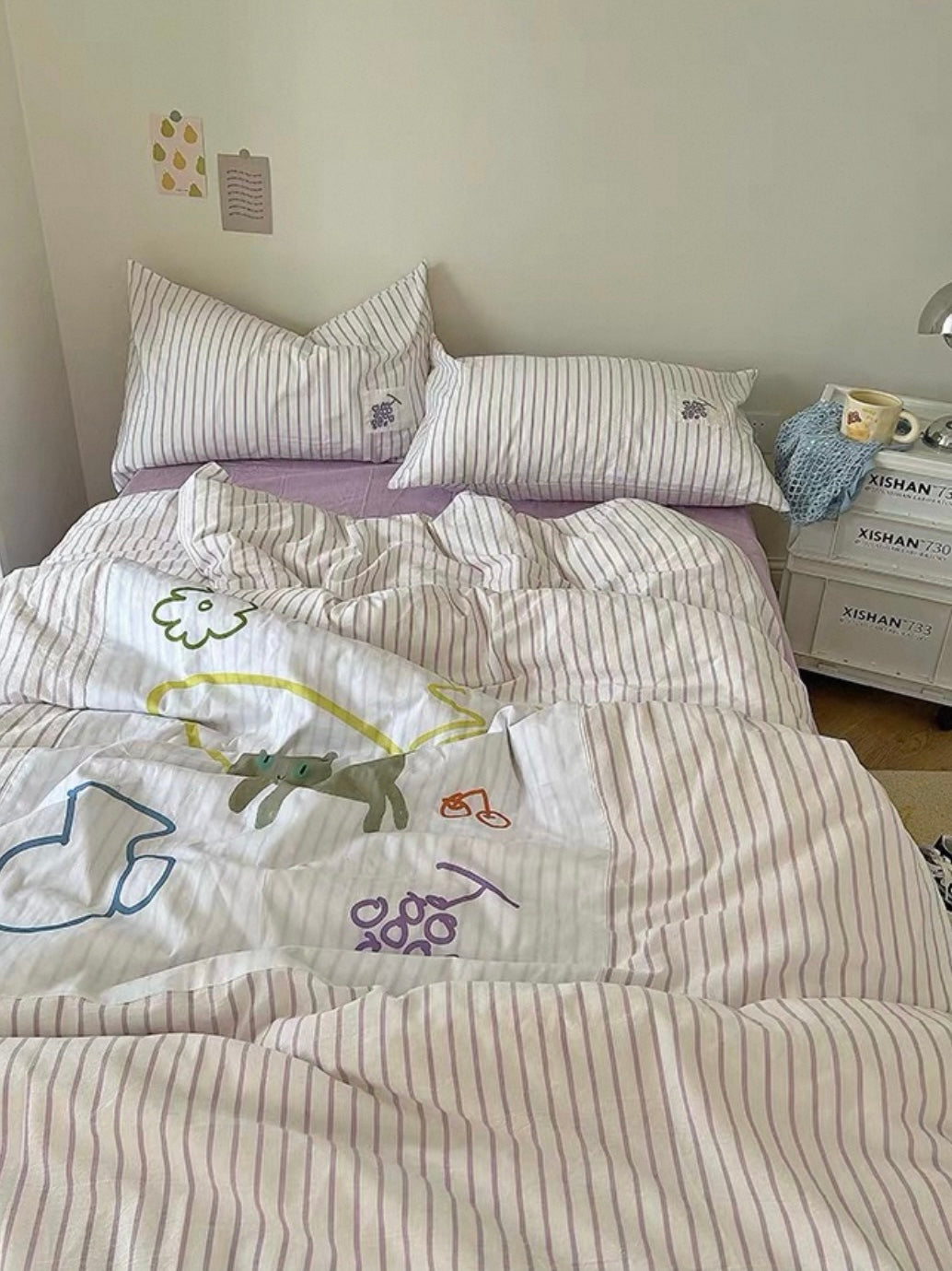 Purple striped bedding set with colorful cartoon patches, matching pillows, and a bedside table.