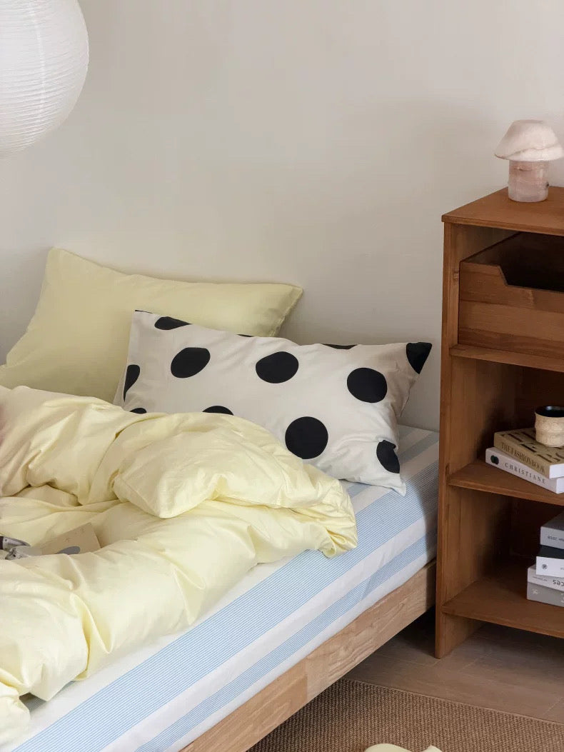 A cozy bedroom corner showing a bed with pale yellow bedding, a white pillow with black polka dots, a light blue striped sheet, and a wooden nightstand beside it.