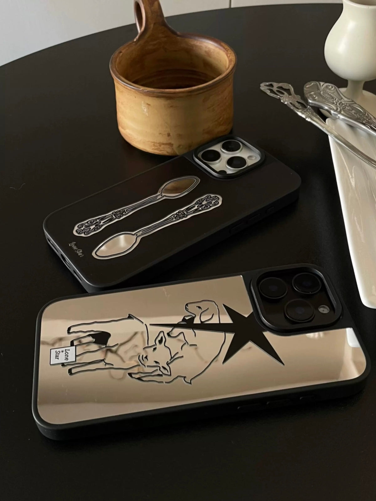 Two reflective phone cases lie at different angles, showing the silver lamb and star print and black spoon print.