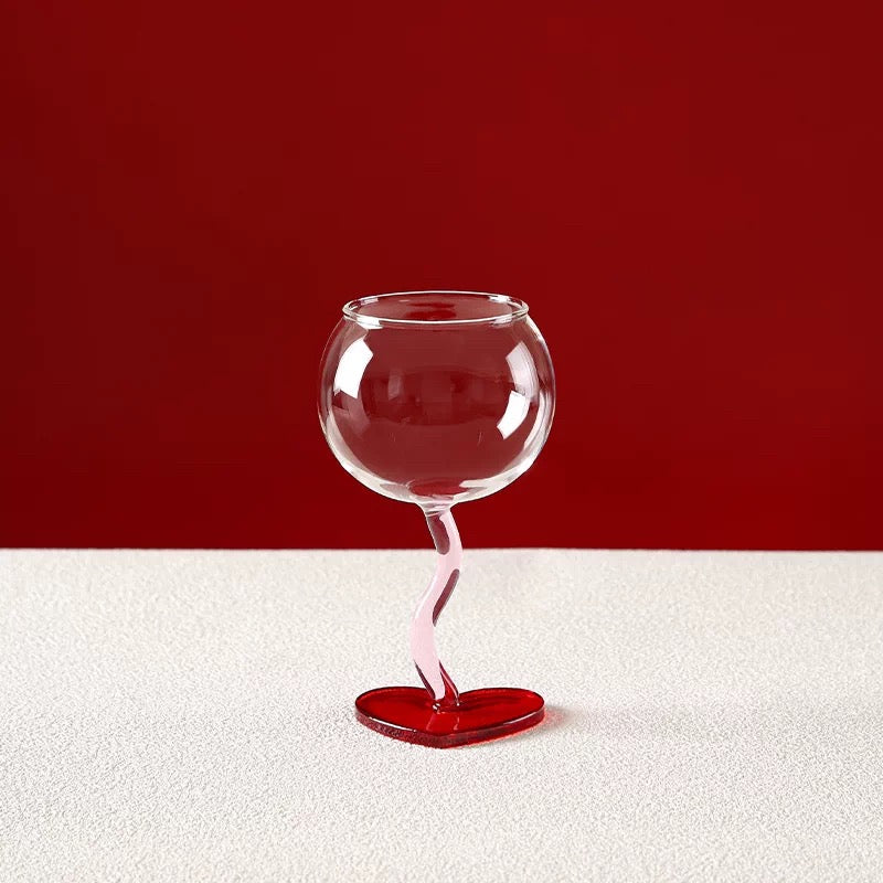 Clear glass wine glass with a red heart-shaped base on a white surface with a red background