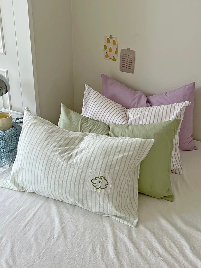 Sage striped pillowcase with a small embroidered flower against green and lilac pillows near a window.