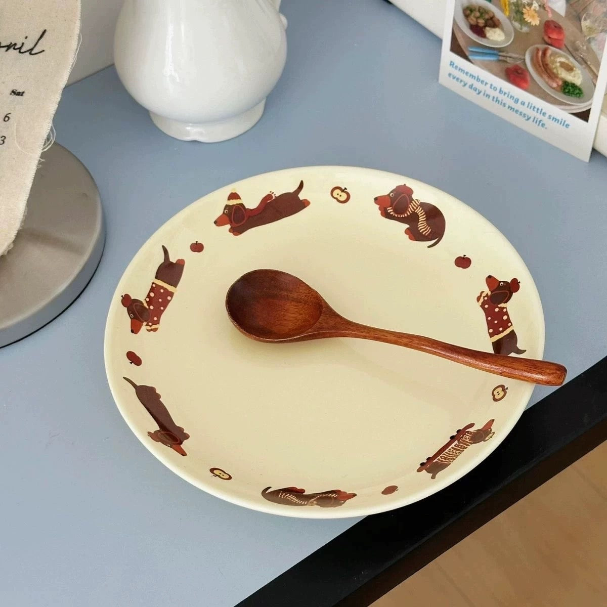A round cream-colored ceramic plate with brown dachshund illustrations, shown on a light blue tabletop with a wooden spoon resting on it.