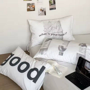 A styled bed setup featuring three white printed pillows with black text and graphic designs, alongside a black laptop on a light wood floor against a white wall.