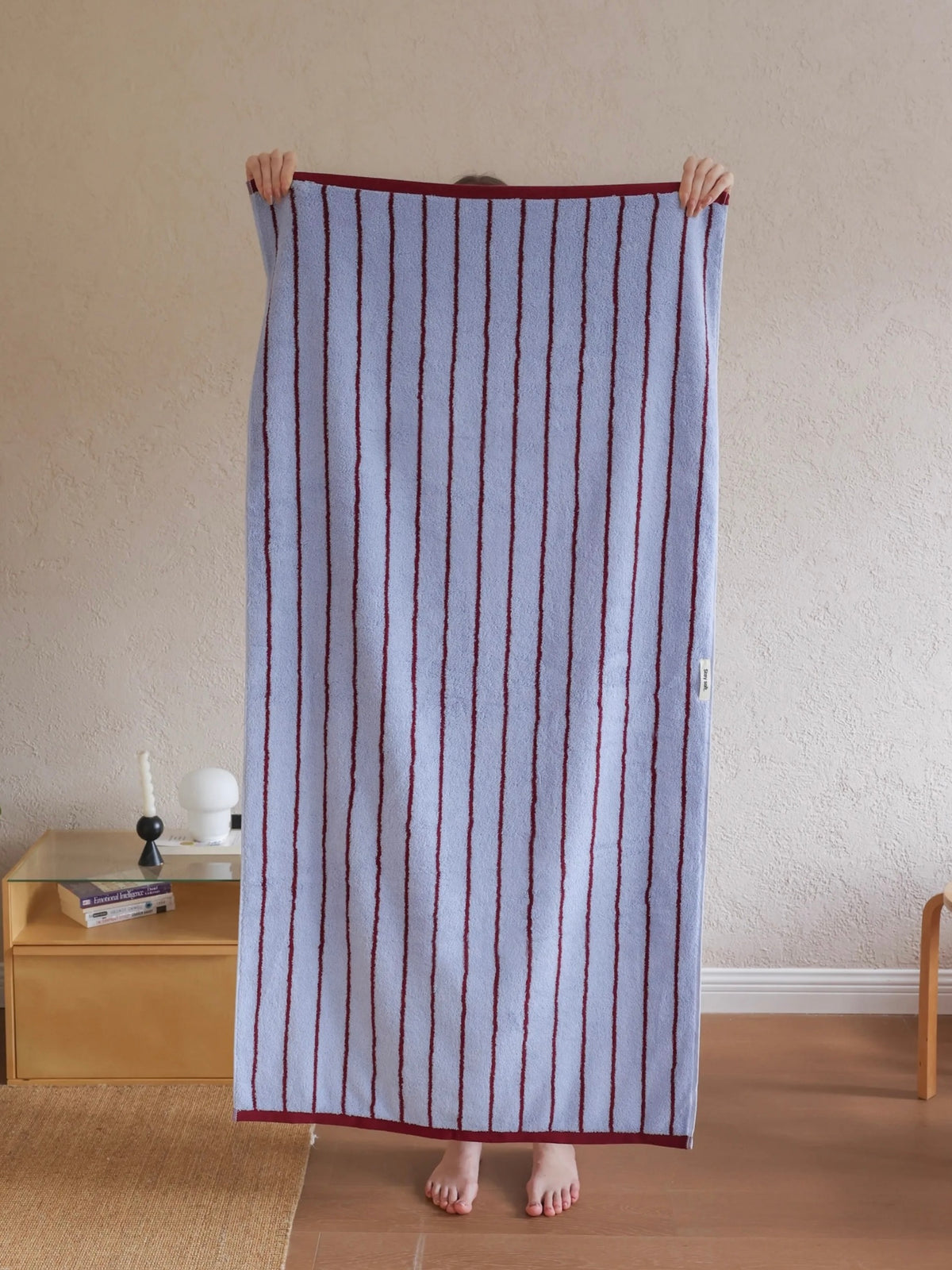 A person holds up a large striped towel or blanket in a home setting. The textile has vertical stripes in dark red/burgundy on a pale lavender-blue background, with a solid dark red border.