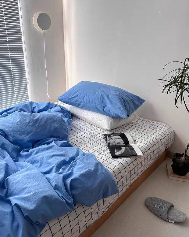 Bed with blue bedding and pillows in a room with a plant and slippers.