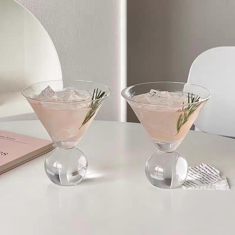 Clear glass with pink cocktail and ice cubes on a white surface with a book underneath.