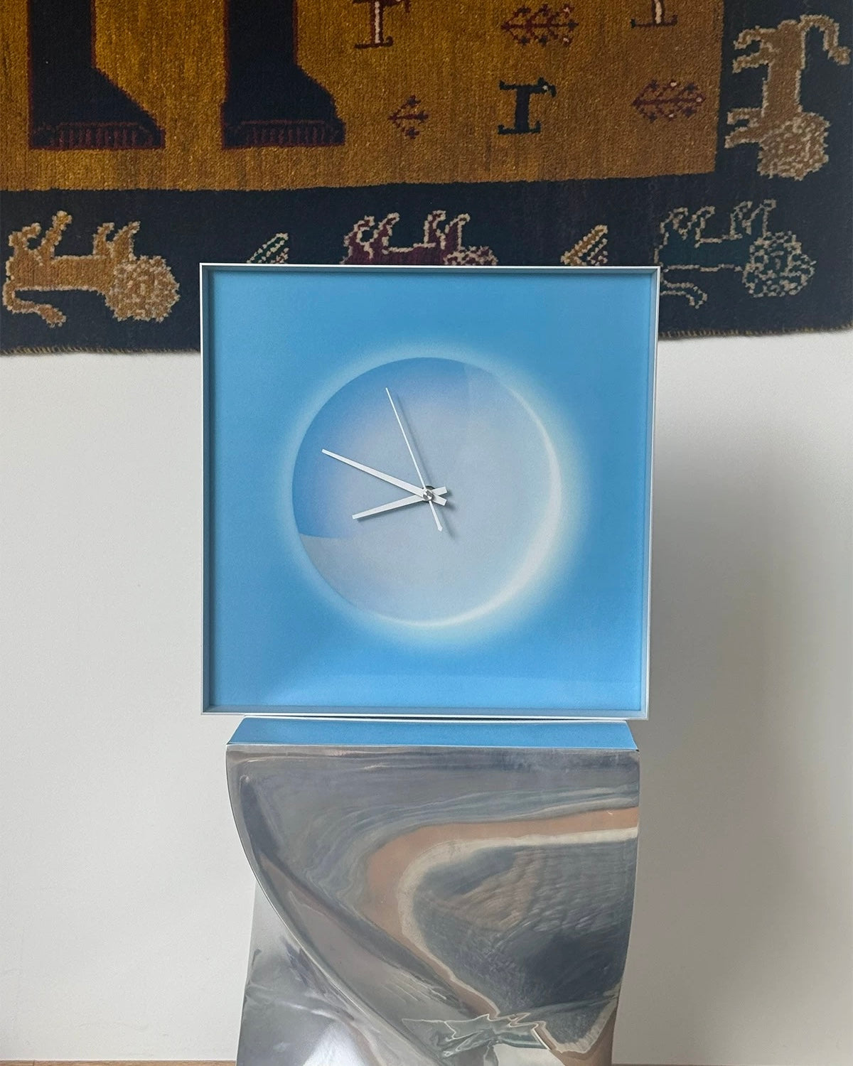 A modern square wall clock with a light blue face and white clock hands, mounted on a reflective metallic stand against a white wall with a patterned rug in the background.