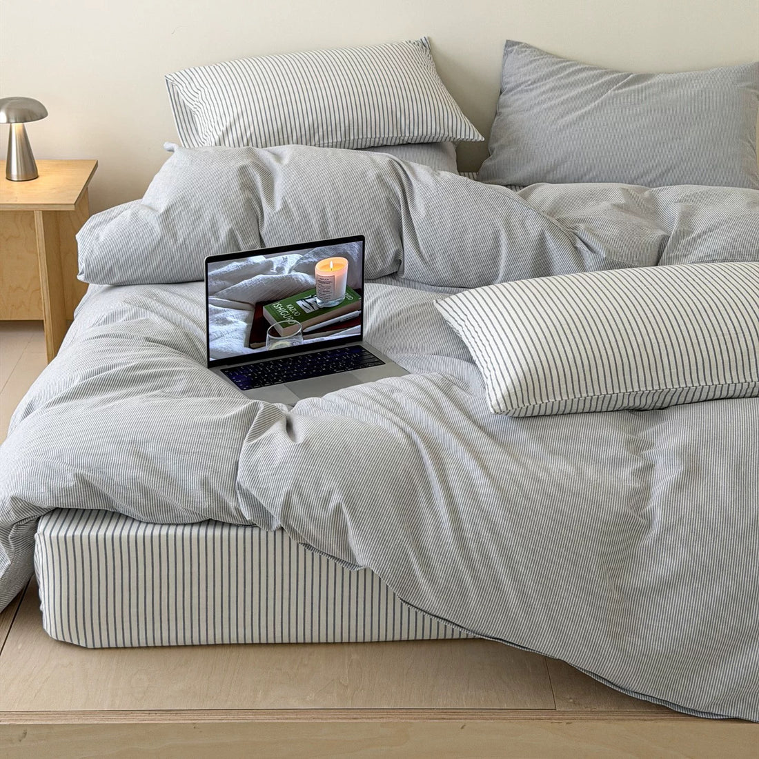A minimalist bedroom scene featuring a light gray and white striped bedding set on a low wooden platform bed. The bedding includes a duvet cover, multiple pillows, and a matching striped mattress protector.