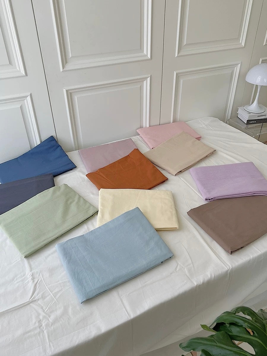 Folded solid color bed sheet options in blue, pink, green and cream are arranged across a white bed.