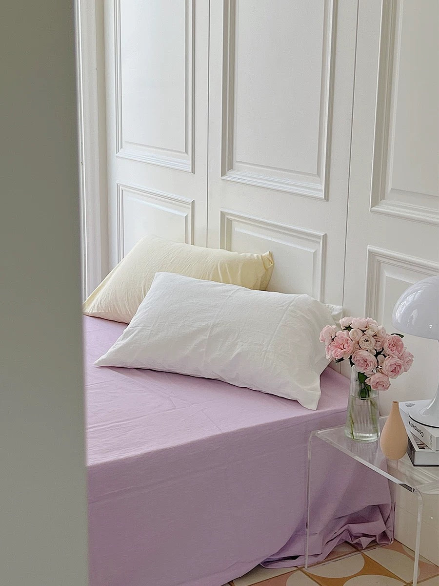 A light purple sheet covers the bed beneath pale yellow and white pillows beside pink flowers.