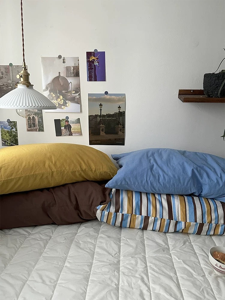 Solid mustard and blue cotton pillowcases are stacked above mocha and striped pillowcases.