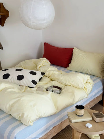 A cozy bedroom scene with a bed made up in pale yellow bedding, a blue-and-white striped sheet, a red pillow, a cream pillow, and a black-and-white polka dot accent pillow. A small round side table with a mug, book, and earbuds sits beside the bed.