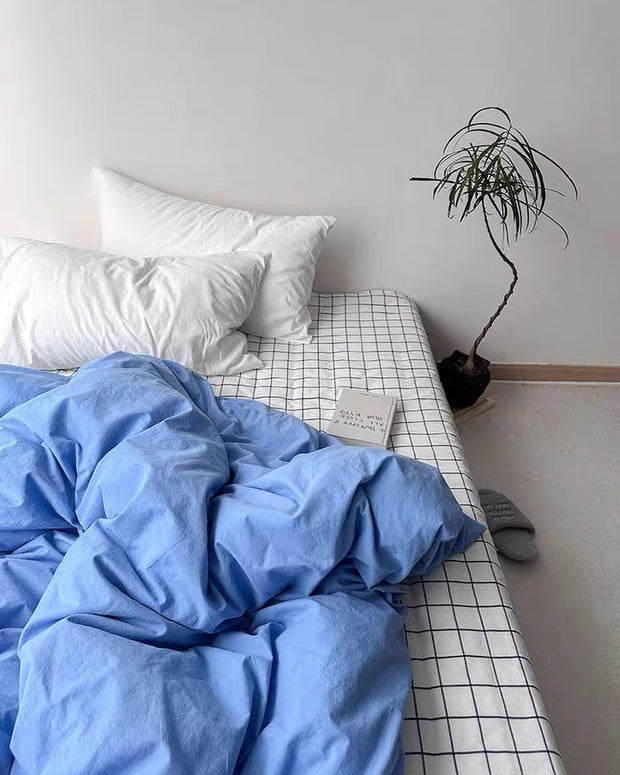 Bed with blue comforter and white pillows, grid-patterned bedspread, and small plant on a shelf.