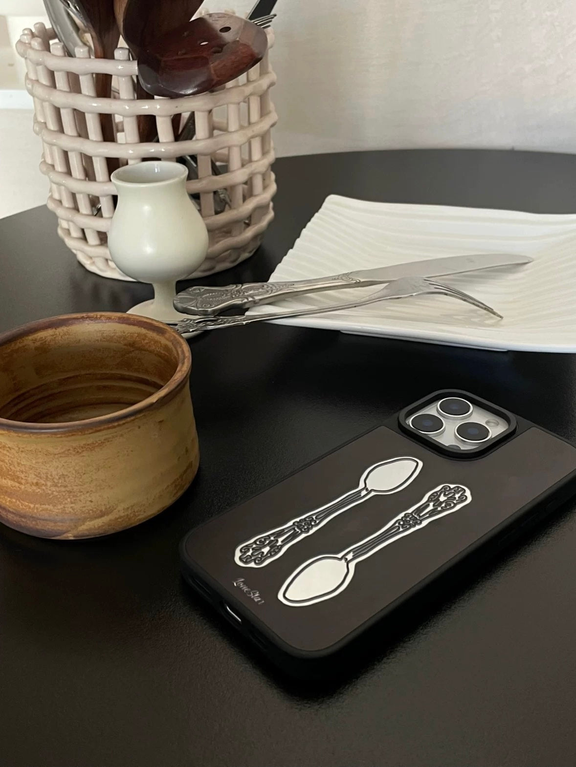 A spoon phone case lies at an angle, showing two ornate spoons facing opposite ways on a black back.