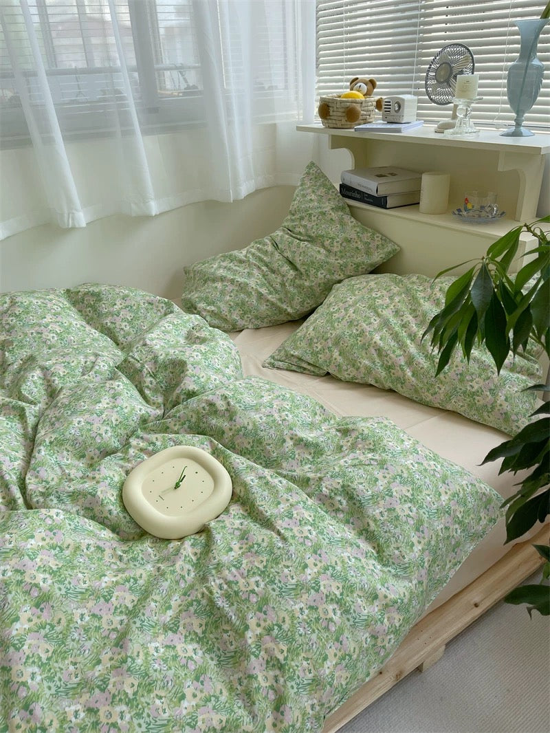 Bed with green floral bedding in a bright room with white curtains and a plant.