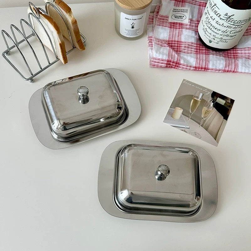 Two silver butter dishes on a white surface with a bottle of wine and a magazine in the background.