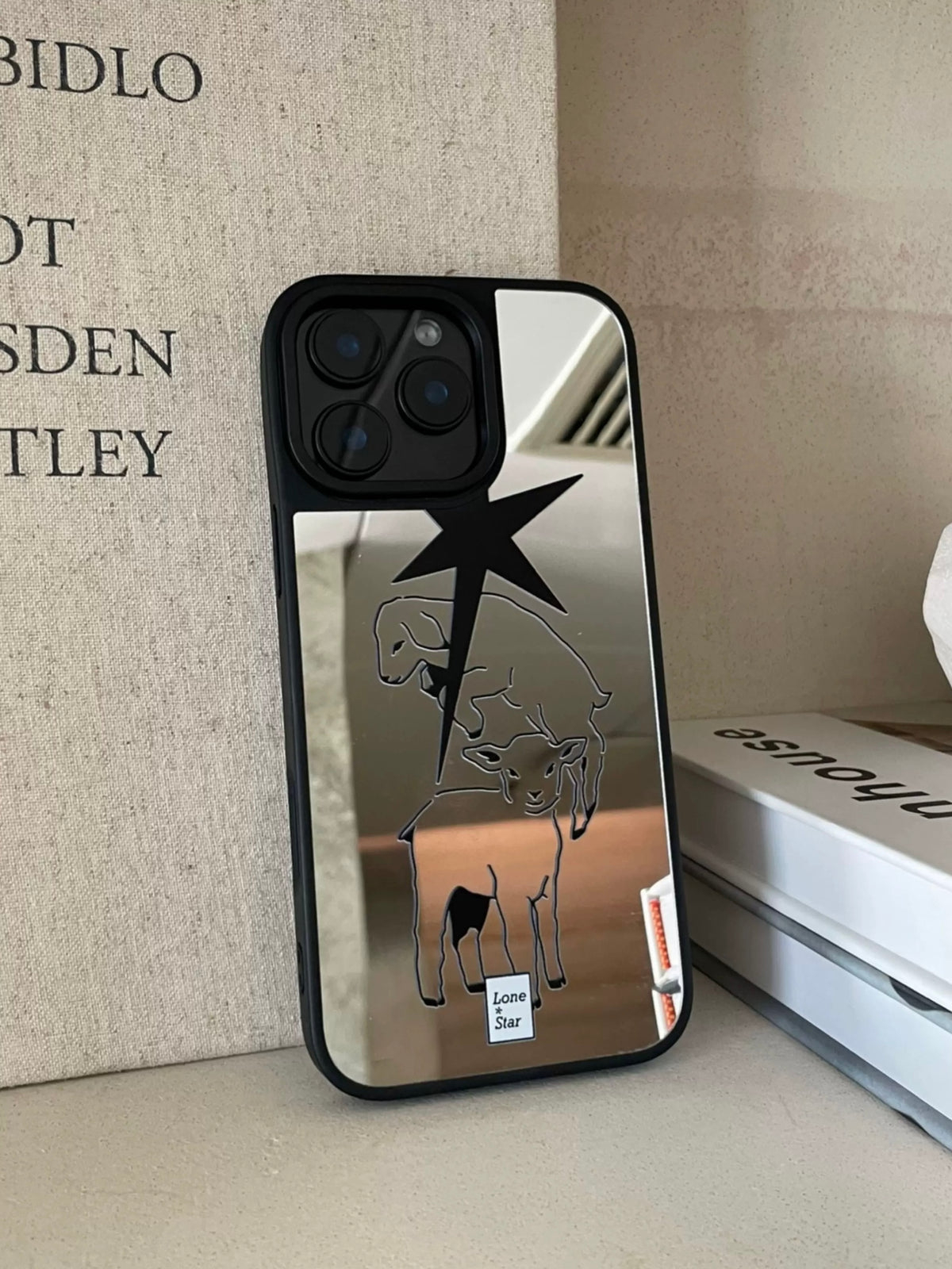 A star phone case stands upright, showing a large black pointed star, lamb outlines and a reflective silver back.