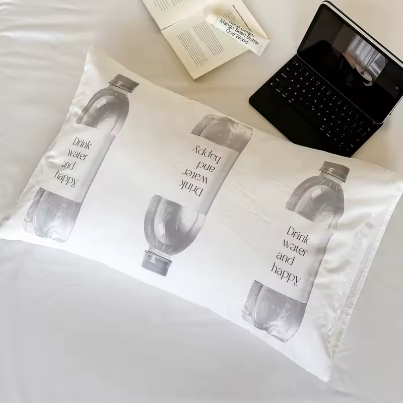A white pillowcase featuring a repeating grayscale print of a water bottle with the words 'Drink water and happy' laid flat on a white bed, with a laptop, book, and phone nearby as styled props.
